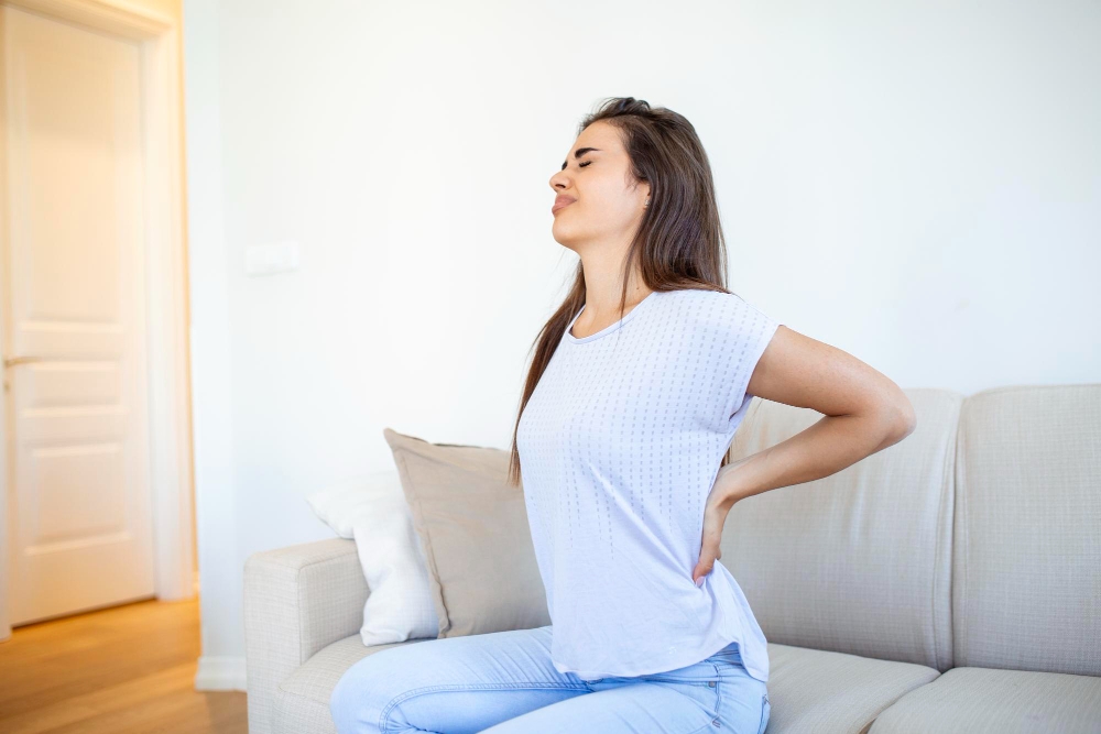 Woman experiencing lower back pain after sleeping while sitting on a couch at home